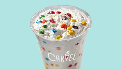 A frozen dessert with whipped cream and multicolored candy sprinkles on top, served in a disposable cup, set against a pale blue background.